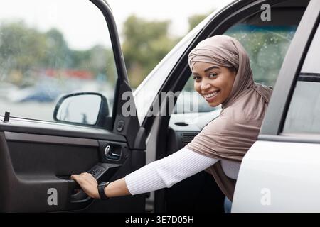 Cheerful Black woman in hijab holding car door sitting on driver seat in city during daytime Stock Photo