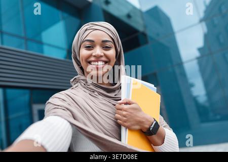 Cheerful Black female student in hijab holding notebooks taking selfie while standing in campus during daytime Stock Photo