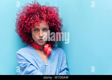 Hispanic woman with red curly hair wearing headphones around her neck pouting her lips while sitting against blue wall Stock Photo