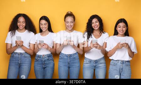 Diverse group of women wearing white shirts and jeans using smartphones standing against yellow background in studio Stock Photo