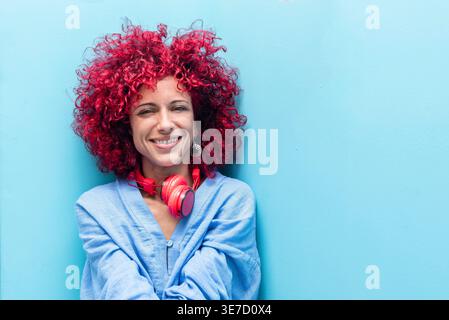 Hispanic woman with red curly hair wearing headphones around her neck smiling while sitting against blue wall Stock Photo