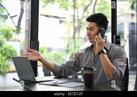 Asian male having phone call looking at laptop while sitting at table with cup of coffee in coffee shop Stock Photo