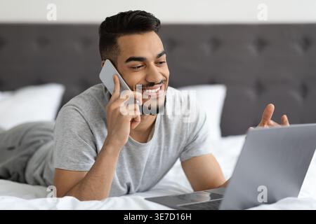 Arab man having phone call and using laptop while lying down on bed in bedroom at home Stock Photo