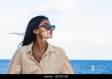 Indian woman with long black hair wearing sunglasses and beige dress looking sideways standing against sea during daytime Stock Photo