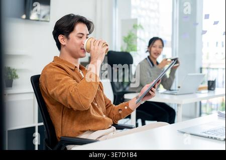 Asian businessman drinking coffee holding clipboard while sitting at table near female colleague in office Stock Photo