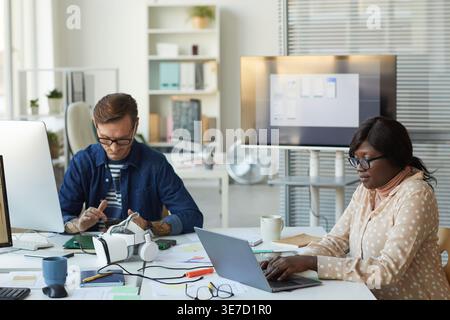 Black female programmer wearing eyeglasses working on laptop near caucasian male colleague while sitting together at desk with computer and virtual... Stock Photo