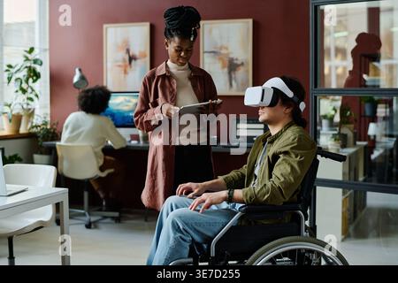 Black female office worker with braids using tablet standing near disabled caucasian male colleague wearing VR headset experiencing virtual reality... Stock Photo