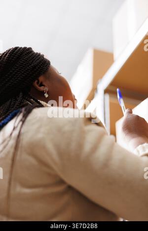 Storehouse worker standing near parcel shelf and pointing upwards with pen. Industrial warehouse african american manager supervising goods storage and dispatching and taking notes Stock Photo