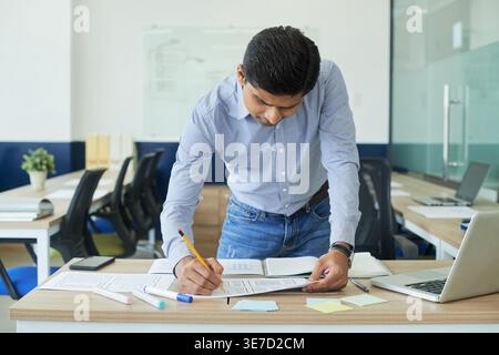 Indian male designer making corrections on webpage layout document with pencil while leaning on desk with laptop and smLifestylephone in office Stock Photo