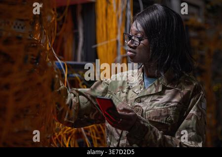 Black female soldier wearing eyeglasses holding smartphone checking cables and wires while standing in server room Stock Photo