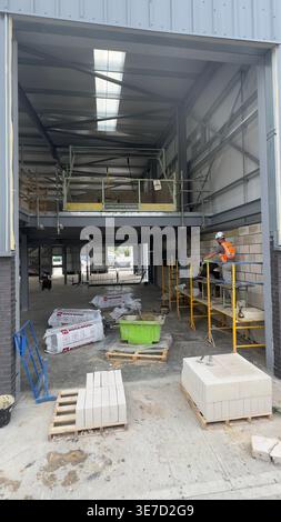 Worker in orange vest stands on yellow scaffolding inside large warehouse with stacked bricks and construction materials. Stock Photo