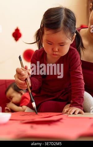 Asian mother and daughter writing with ink and brush doing traditional calligraphy while sitting on sofa with baby girl in living room at home Stock Photo