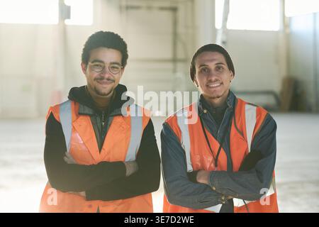Mixed-race male workers wearing safety vests with arms crossed standing in warehouse Stock Photo