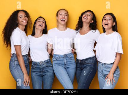 Cheerful diverse group of women wearing white shirts and jeans laughing while standing together against yellow background in studio Stock Photo