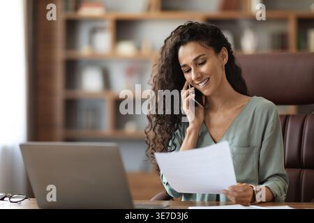 Middle Eastern woman with curly hair holding paper having phone call while sitting at table in office Stock Photo