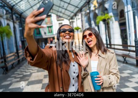Black and caucasian female friends wearing trench coats and sunglasses taking selfies using smLifestylephone while standing in city during daytime Stock Photo