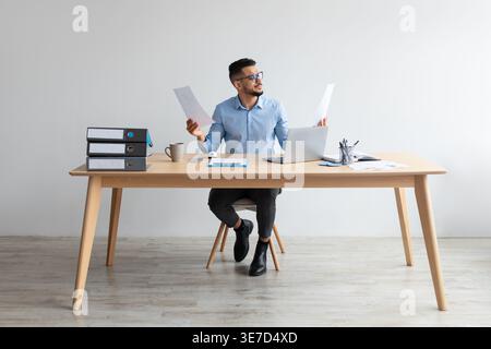Middle Eastern man wearing eyeglasses holding documents sitting at table with laptop in office Stock Photo