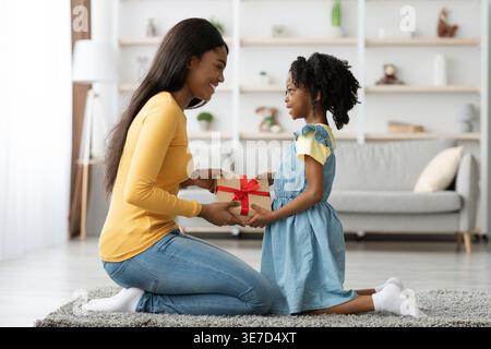 Black woman receiving gift box from daughter kneeling on floor carpet in living room at home Stock Photo