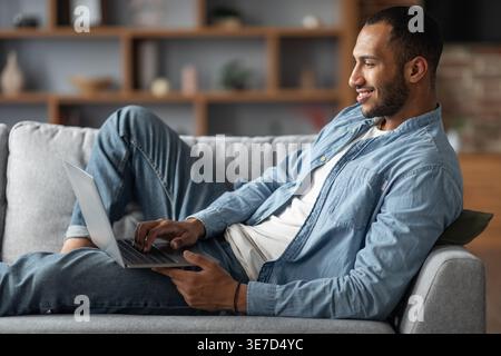 Smiling Black man working on laptop lying on sofa in living room at home Stock Photo