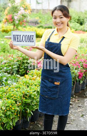 Smiling Asian female worker holding welcome signage while standing in plant market Stock Photo
