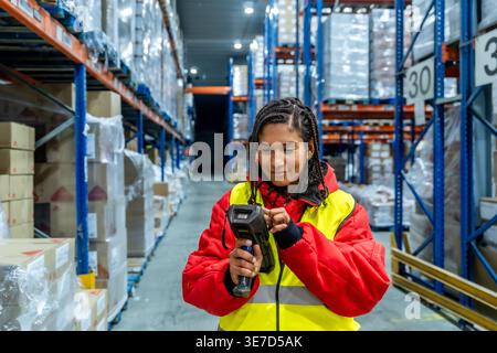 Hispanic female worker with braids wearing red jacket and safety vest operating barcode scanner while standing in cold storage warehouse Stock Photo