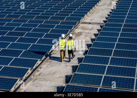 Hispanic and caucasian female engineers wearing hard hats and safety vests discussing work while walking on commercial rooftop with solar panels on... Stock Photo