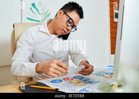 Asian male graphic designer rearranging icons while using smartphone sitting on chair in office Stock Photo