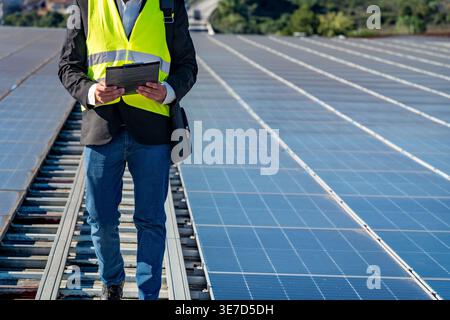 Cropped image of Hispanic male engineer in safety vest holding clipboard standing on rooftop of solar panels on sunny day Stock Photo