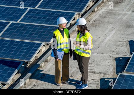Hispanic and caucasian female engineers wearing hard hats and safety vests discussing work while walking on commercial rooftop with solar panels on... Stock Photo