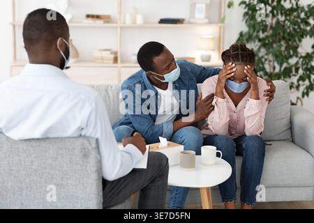 Black man comforting his wife having mental health problems while sitting on couch and talking with male therapist in office during covid-19 pandemic Stock Photo