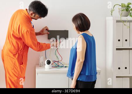 Indian male technician showing video surveillance equipment to senior woman while standing in house of customer Stock Photo