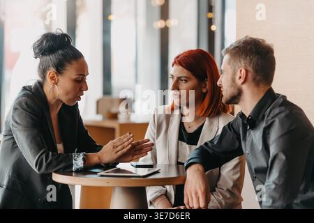 Serious Hispanic business woman discussing business plan with caucasian male and female colleagues while sitting together at table in the office Stock Photo