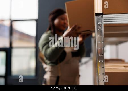 Selective focus of metallic shelves full with carton boxes, african american worker preparing customers order in storage room. Stockroom employee wearing industrial overall working at storehouse Stock Photo