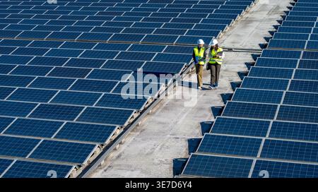 Hispanic and caucasian female engineers wearing hard hats and safety vests discussing work while walking on commercial rooftop with solar panels on... Stock Photo