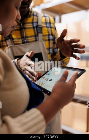 Inventory analyst explaining warehouse employee data on tablet screen. African american industrial storehouse manager holding portable device and analyzing stock supply analytics Stock Photo