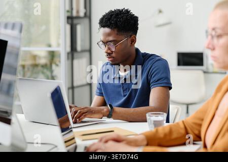 Serious Black male programmer in eyeglasses sitting at desk with caucasian female colleague developing new software on computers working together i... Stock Photo
