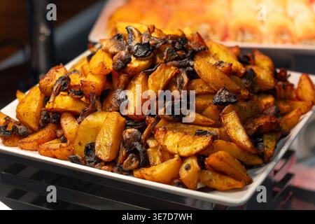 Close-up of delicious roasted potato wedges with mushrooms on a white platter, served at a hotel buffet or restaurant with warm lighting and blurred b Stock Photo