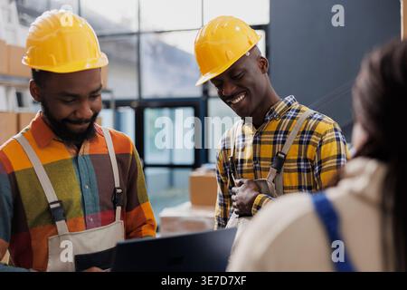 Warehouse workers wearing yellow helmets laughing and chatting at work. Smiling african american men storehouse employees in protective hard hats and overall having fun conversation Stock Photo