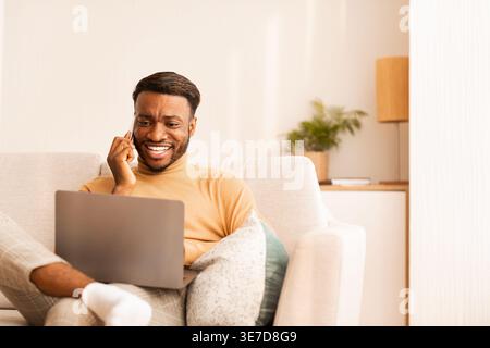 Black man looking at laptop having phone call while sitting with crossed legs on sofa in living room at home Stock Photo