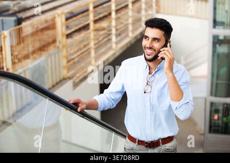 Smiling Middle Eastern man having phone call looking away standing on escalator in city during daytime Stock Photo