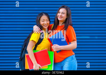 Smiling Asian female student with backpack holding notebooks and folder standing with caucasian female friend against blue wall on university campu... Stock Photo