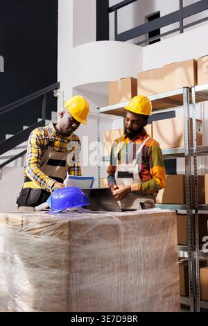 Logistics managers analyzing inventory and comparing data on laptop and clipboard in warehouse. African american men checking goods supply schedule and working in distribution department storehouse Stock Photo