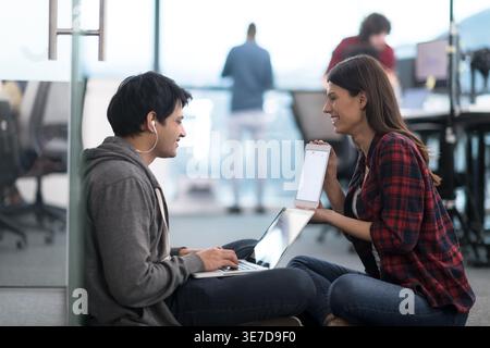 Smiling Mixed race man with earphones using laptop with caucasian female coworker showing tablet sitting on floor in office Stock Photo
