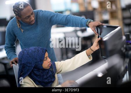 Focused Black man using computer standing behind Black muslim female coworker sitting during meeting in office Stock Photo