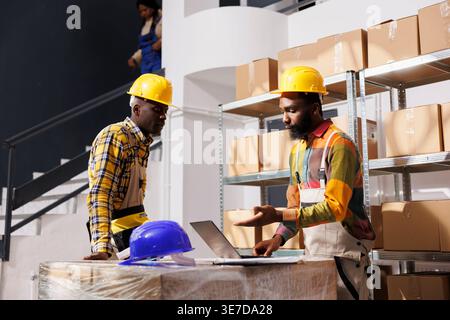African american logistics managers discussing inventory management software while checking goods in stock on laptop. Distribution department employees talking about supply chain in storehouse Stock Photo