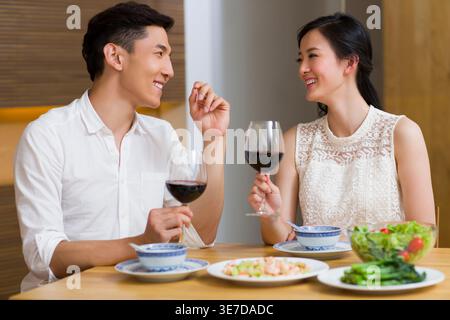 Cheerful Chinese couple holding glass of wine having a dinner while sitting at table at home Stock Photo