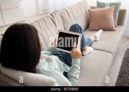 Indian female using tablet lying on sofa in living room at home Stock Photo