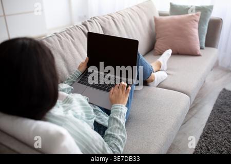 Indian female using laptop lying on sofa in living room at home Stock Photo