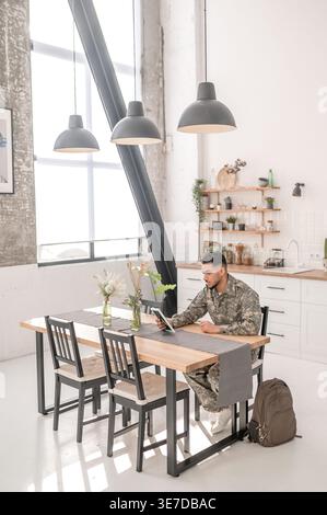 Arab military man with sad facial expression wearing camouflage uniform holding and looking at picture frame while sitting at kitchen table at home Stock Photo