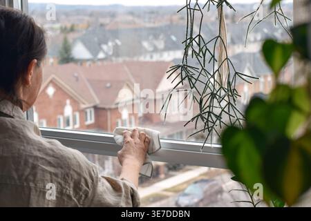 Woman cleaning window frame in apartment using rag. Indoor home cleaning scene focusing on household chores, surface wiping, and maintaining tidy livi Stock Photo